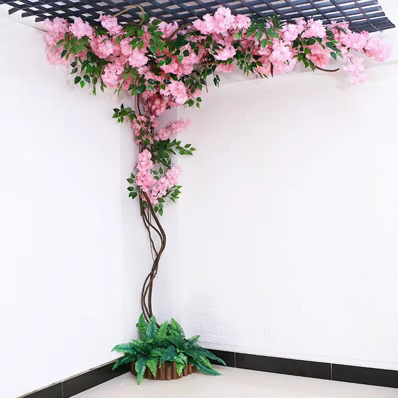 Artificial pink cherry blossom vine climbing a corner wall and ceiling, realistic silk flowers with green leaves and a decorative fern base.