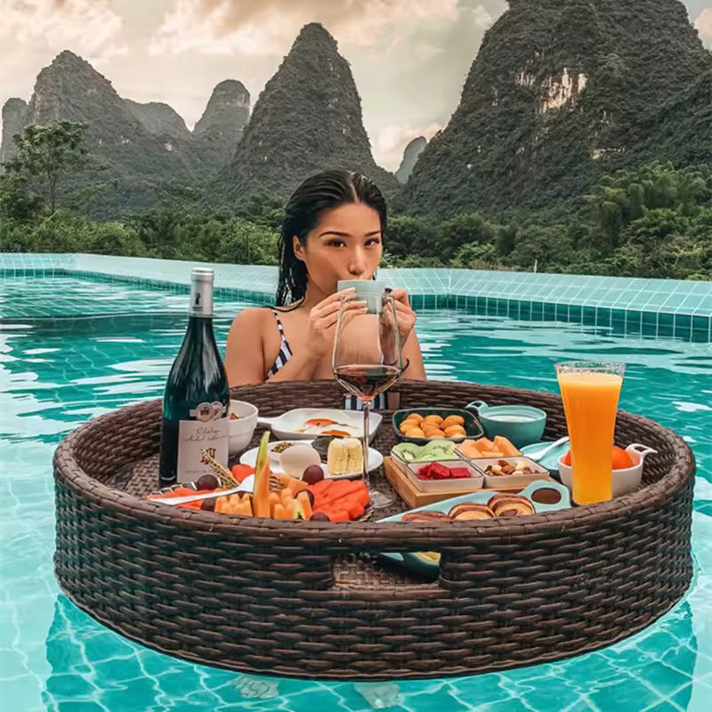 Luxury round dark brown floating rattan tray in a turquoise pool, filled with wine, juice, fruit, and breakfast dishes against a mountain backdrop.