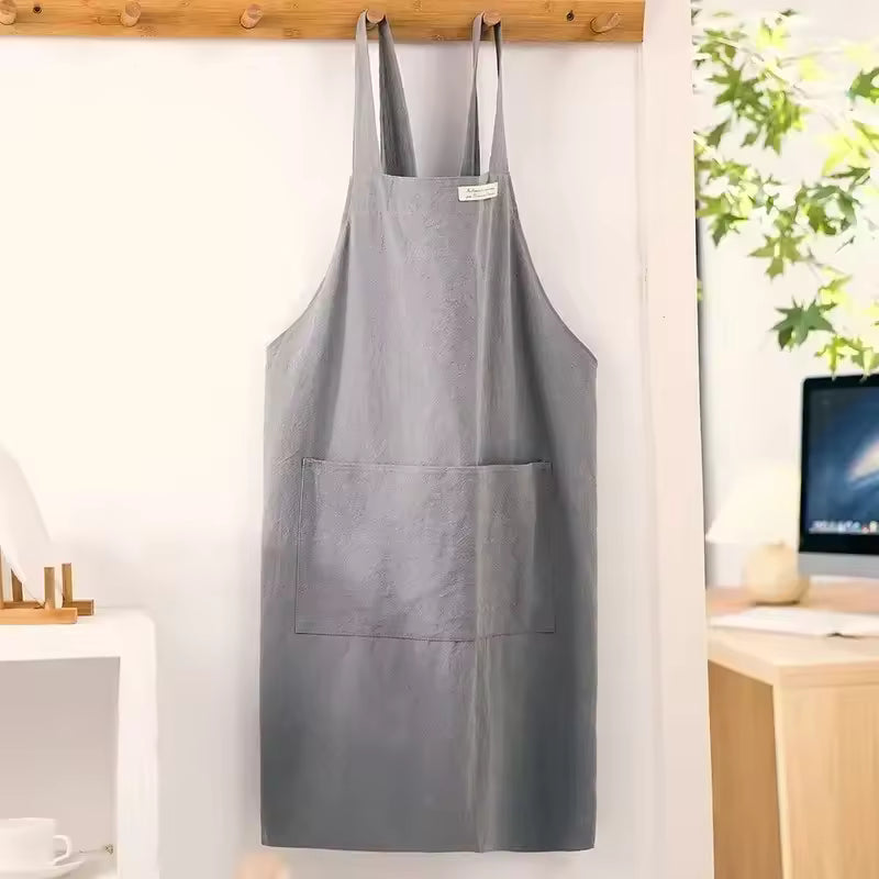 Grey minimalist cotton-linen apron with cross-back straps and a large front pocket hanging on a wooden wall hook.