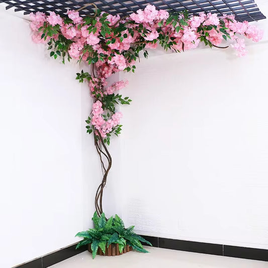 Artificial pink cherry blossom vine climbing a corner wall and ceiling, realistic silk flowers with green leaves and a decorative fern base.