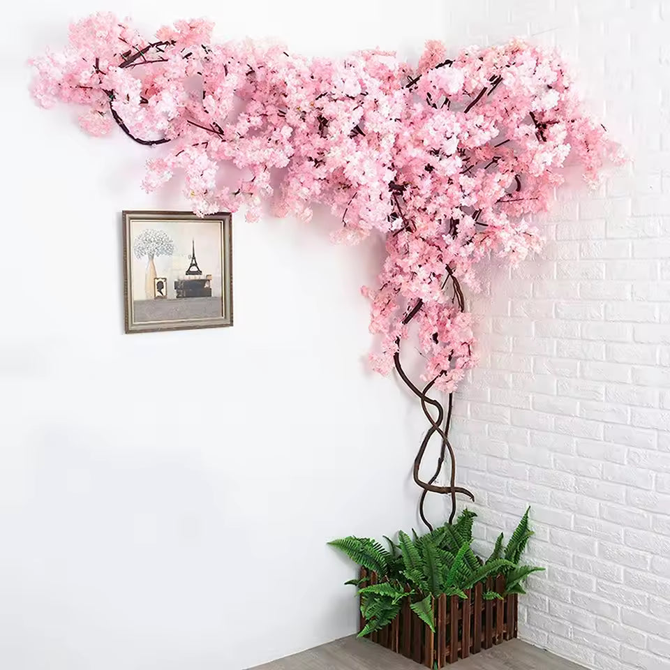 Corner wall decoration with artificial pink cherry blossoms and brown branches, growing from a wooden planter with green ferns against a white brick wall.
