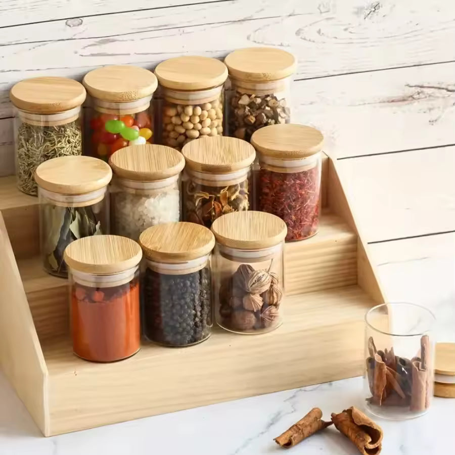 Set of 12 clear glass storage jars with airtight bamboo lids, filled with various spices and condiments on a wooden display stand.