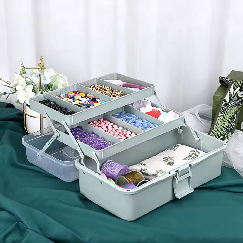 Sage green 3-layer plastic craft organizer box with a clear lid, showing 180-degree cantilever trays filled with colorful beads, pencils, and sewing supplies.