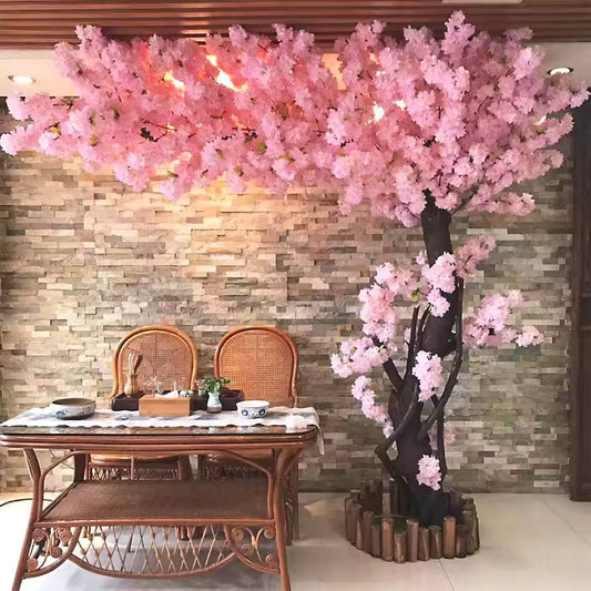 A large, realistic pink artificial cherry blossom tree with a thick trunk and lush silk flowers, standing next to a wooden dining table against a stone wall.