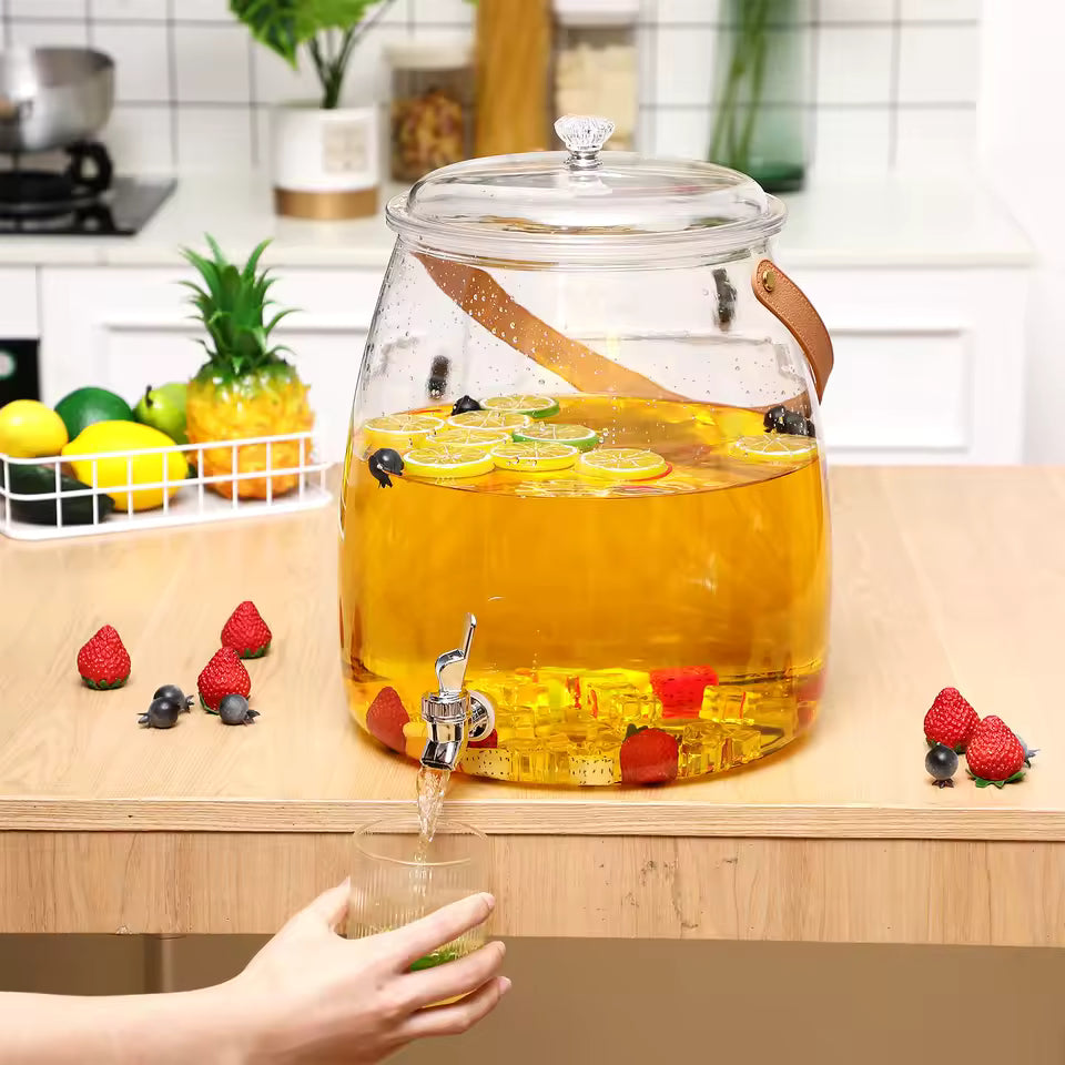 Stylish clear plastic beverage dispenser filled with yellow lemonade and fruit, featuring a stainless steel tap and a brown leather handle.