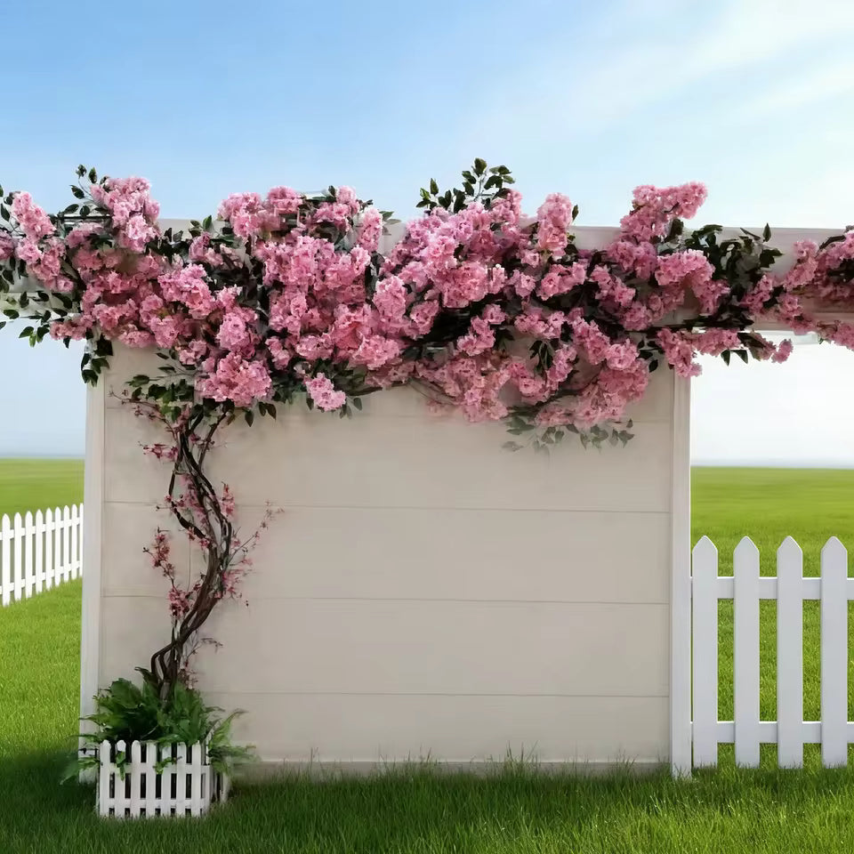 Realistic artificial cherry blossom tree with pink silk flowers climbing along a white wooden wall and fence in an outdoor garden setting.