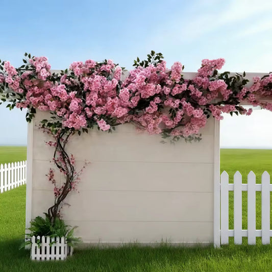 Realistic artificial cherry blossom tree with pink silk flowers climbing along a white wooden wall and fence in an outdoor garden setting.