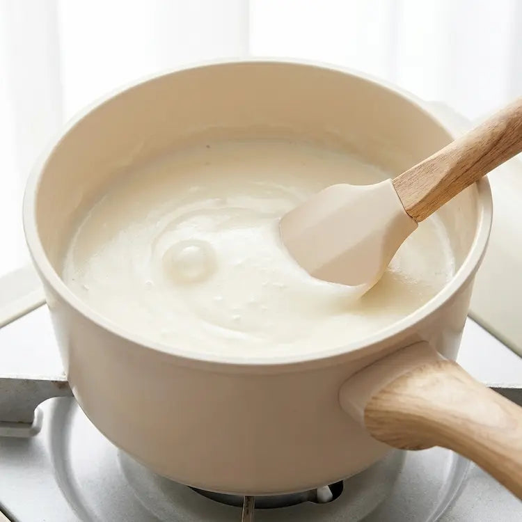 A high-angle close-up of a smooth, creamy white béchamel sauce simmering in a small cream ceramic saucepan. A matching cream silicone spatula with a wood handle is used to stir the sauce, showing no signs of sticking or scorching on the non-stick surface. The image highlights the superior heat distribution and easy cleanup of the PFOA-free ceramic coating. Clean, bright kitchen aesthetic from Nova Daily Shop.
