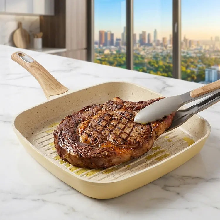A professional high-key culinary close-up shot of the square cream-colored ceramic nonstick grill pan from the 26-piece collection, positioned on a sleek black induction cooktop. A large, perfectly seasoned ribeye steak is being seared, displaying distinct, parallel dark grill marks and a rich caramelized crust on its surface. Matching grey-and-wood handle silicone tongs are present. Focus is razor-sharp on the appetizing meat texture and the clean ceramic grill ridges, demonstrating high-heat non-stick per