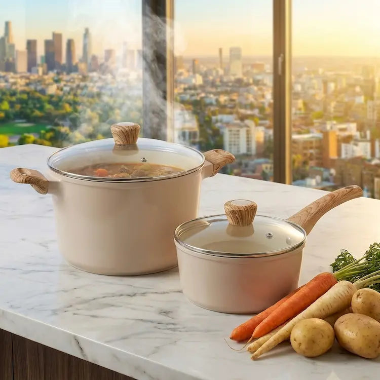 A premium lifestyle shot of a cream-colored ceramic stockpot and saucepan from the 26-piece collection on a white marble counter. The large stockpot is filled with steaming vegetable stew, while fresh carrots, parsnips, and potatoes lie alongside. Both pots feature tempered glass lids and ergonomic wood-look handles. The background shows a luxury kitchen with a sunset city skyline view. Non-toxic, PFOA-free healthy cookware by Nova Daily Shop.