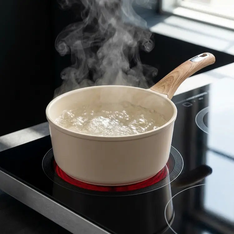 A technical lifestyle shot of a cream-colored ceramic saucepan from the 26-piece set placed on a black glass induction cooktop. The induction ring glows red as water boils vigorously inside the pot, creating thick steam. The image demonstrates the high-performance induction base and heat distribution of the non-toxic, PFOA-free cookware. Modern kitchen setting with soft daylight. Premium quality kitchenware by Nova Daily Shop.