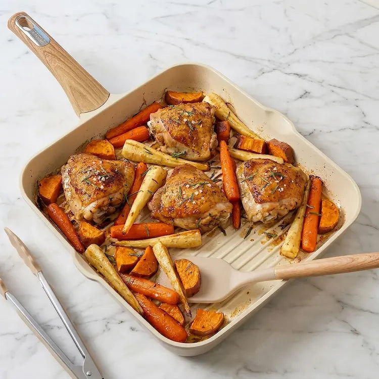 A professional overhead photograph of a square cream-colored ceramic nonstick grill pan with a wood-look handle on a white marble countertop. The pan contains four golden-brown roasted chicken thighs surrounded by caramelized carrots and parsnips, seasoned with sea salt and fresh rosemary. A matching cream silicone spatula rests inside the pan, highlighting the ridged grill surface. Matching silicone tongs are visible to the side. High-end, non-toxic, healthy cooking presentation.
